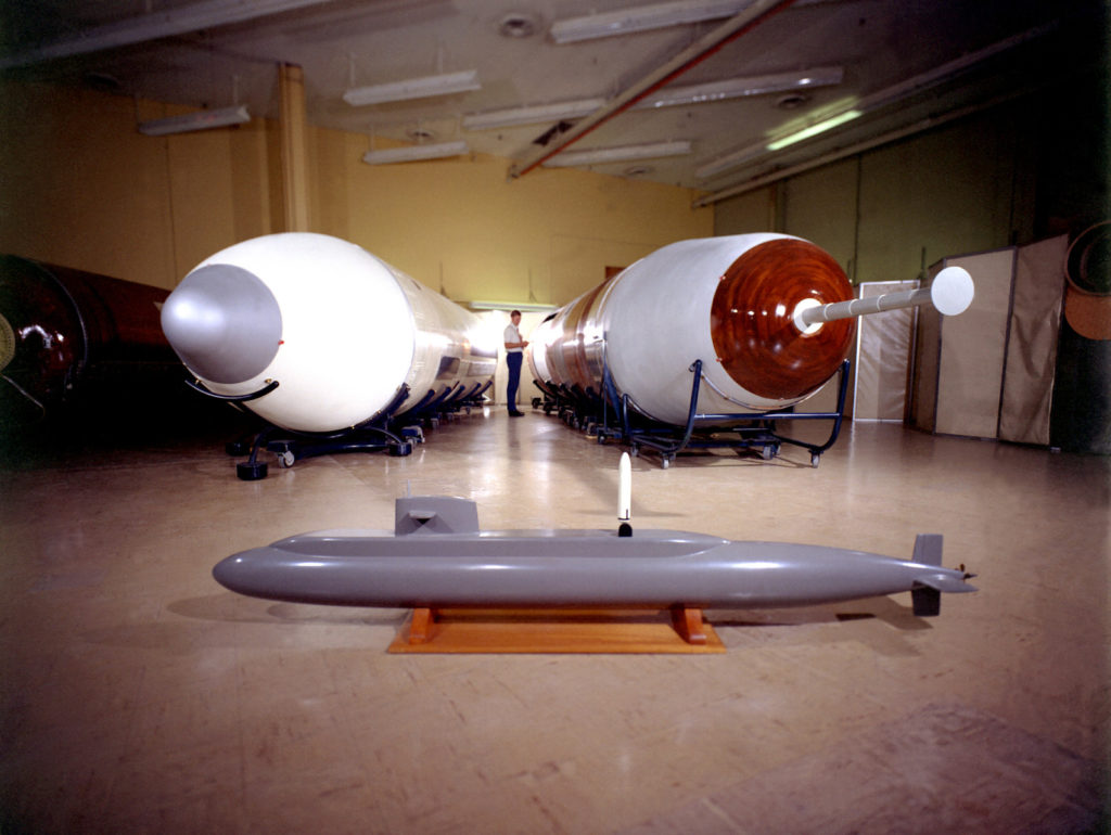A view of a mock-up of a Trident I C-4 missile, right, and its predecessor, the Poseidon C-3 missile.  In the foreground is a model of the nuclear-powered fleet ballistic missile submarine USS ULYSSES S. GRANT (SSBN-631) showing a simulated launching of a Poseidon missile