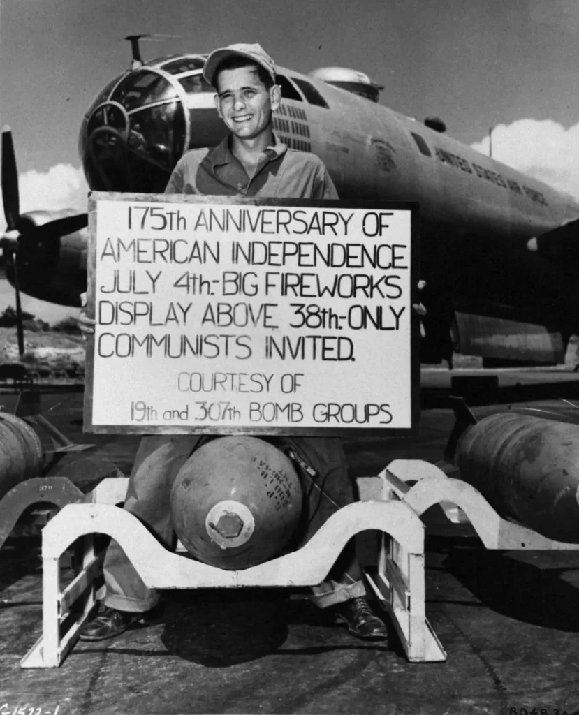 korean war b-29 armament technitian holds a sign displaying the schedule of events while standing over one of the large explosives that will detonate on an enemy target on the 4th of July